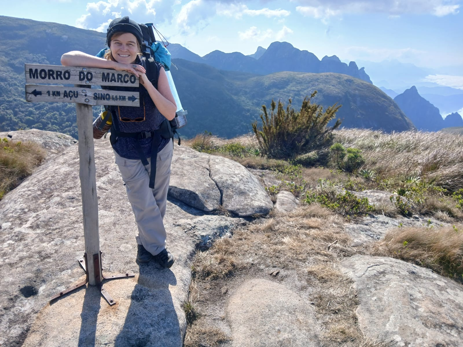 Pessoa sorridente com mochila de trilha está encostada em uma placa de madeira que indica “Morro do Marco – 1 km Açu | 6,5 km Sino”. Ela veste calça clara, camiseta azul e boné preto, e carrega bastões de caminhada presos à mochila. O cenário ao fundo é uma paisagem montanhosa imponente, com picos recortados, vegetação rasteira e céu parcialmente nublado. A luz do sol realça as cores do ambiente natural, transmitindo sensação de tranquilidade e conquista.
