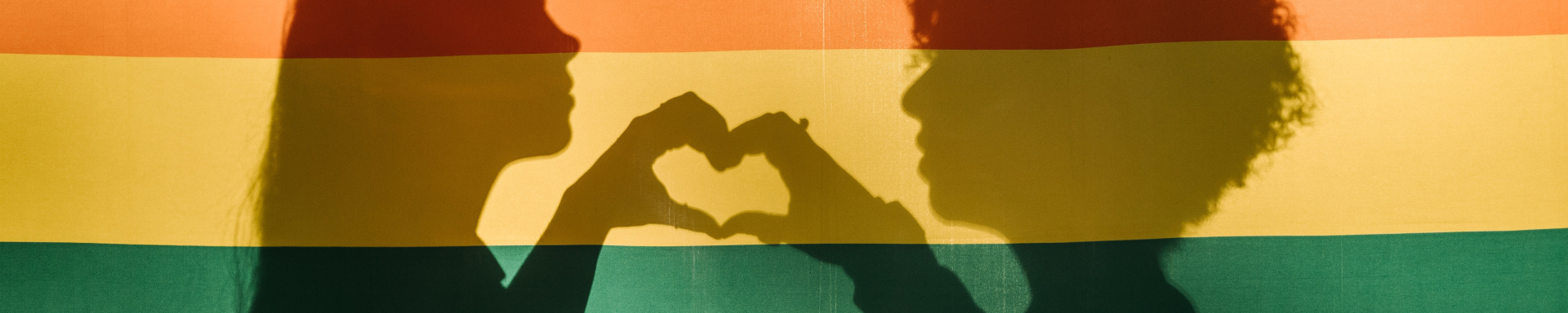 Sombras de duas mulheres fazendo um coração com as mãos situadas em uma bandeira de arco-íris LGBTQIA+