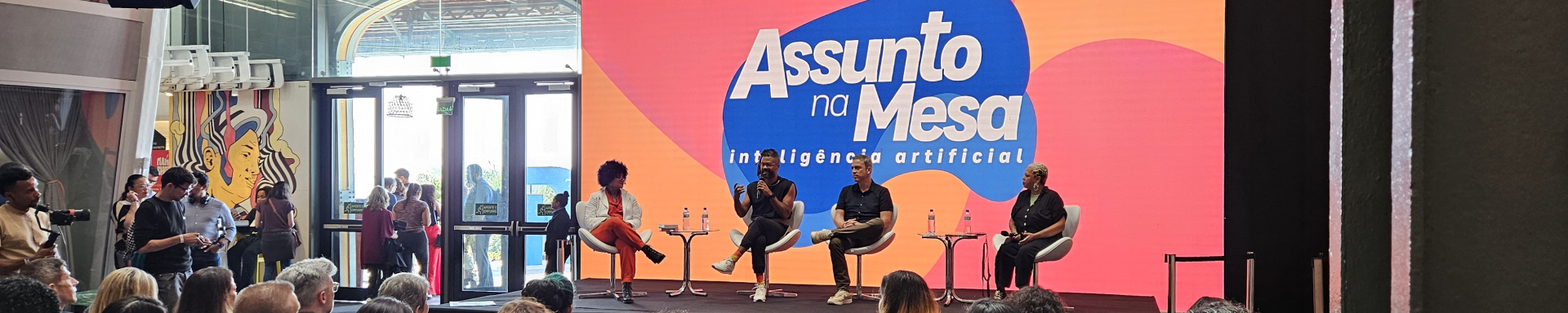 Quatro pessoas estão sentadas em poltronas sobre um palco. Ao fundo, colorido, o nome do programa que abordavam ali: Assunto na Mesa.