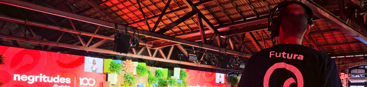 A imagem mostra um auditório coberto, com teto de madeira e vigas aparentes, lotado de pessoas assistindo a um evento. À frente do público, há um grande palco decorado com painéis coloridos e plantas verdes. Um telão vermelho exibe as palavras “negritudes @globo” e “100 anos de Globo” em branco. Há também pequenas telas que mostram a imagem de uma apresentadora.  À direita da imagem, em primeiro plano, um cinegrafista do Canal Futura está de costas, operando uma câmera sobre um tripé. Ele veste uma camisa p